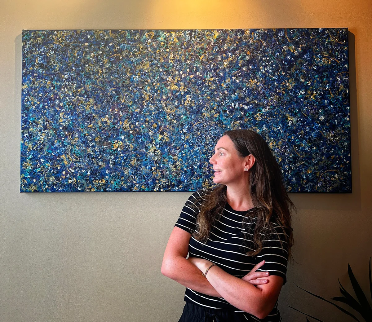 Kelly standing in front of Beginning, a large blue, turquoise, gold, white, and purple abstract painting displayed on a wall.
