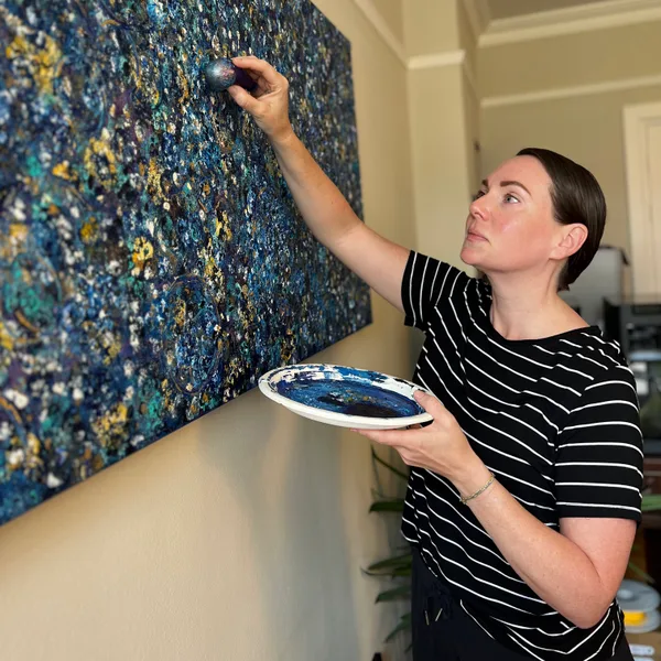 Kelly applying paint to a large blue, teal, and gold abstract artwork.