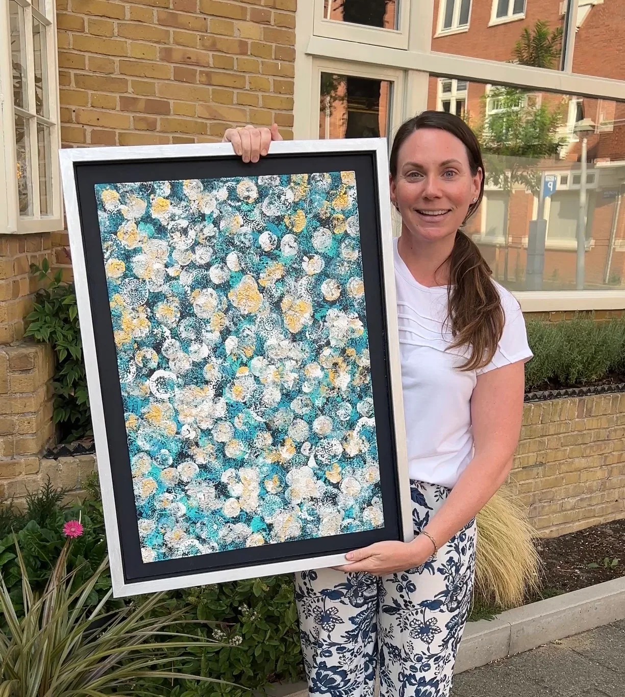 Kelly holding Midnight & Magnolias, a framed turquoise, black, white, and gold abstract floral painting.